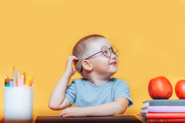 Boy in round glasses in a shirt and sitting at desk and thinking, doing homework, on a yellow background. back to school — Stock Photo, Image