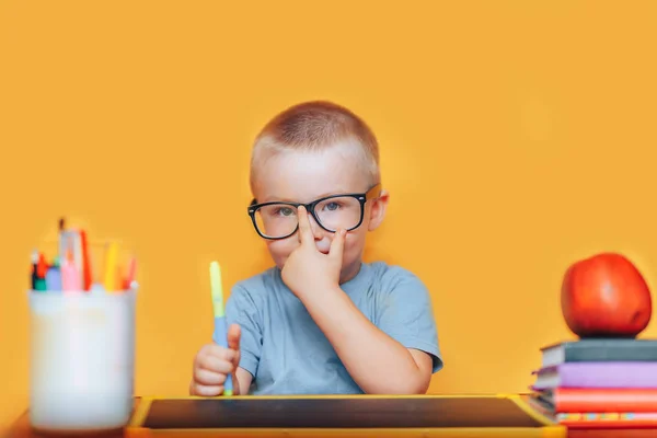 행복 금발의 영리한 소년은 안경에 책상에 앉아 웃고있다. 학교에 갈 준비가 되었습니다. 학교로 돌아갑니다. 책상위에 있는 사과, 펜, 책 — 스톡 사진