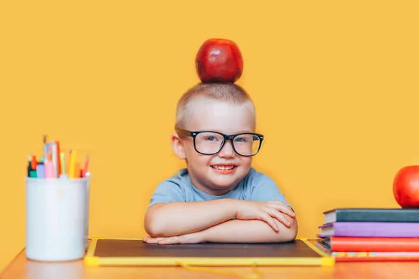 행복 귀여운 영리한 소년은 책상에 앉아 미소. 아이가 노란색 배경에 있습니다. 학교에 처음. 학교로 돌아갑니다. 그의 머리에 사과와 안경에 동공 — 스톡 사진