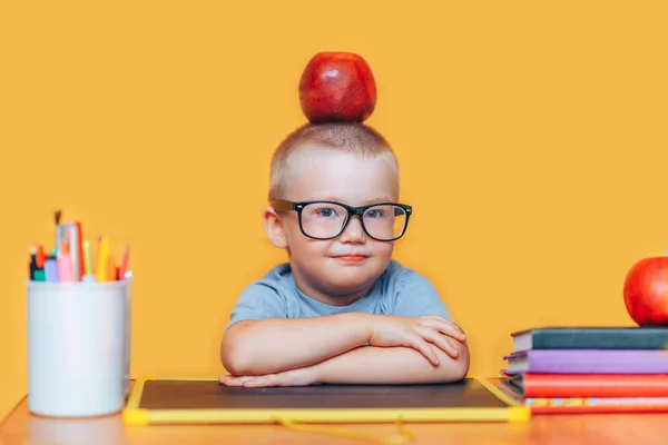 행복 귀여운 영리한 소년은 책상에 앉아 미소. 아이가 노란색 배경에 있습니다. 학교에 처음. 학교로 돌아갑니다. 그의 머리에 사과와 안경에 동공 — 스톡 사진