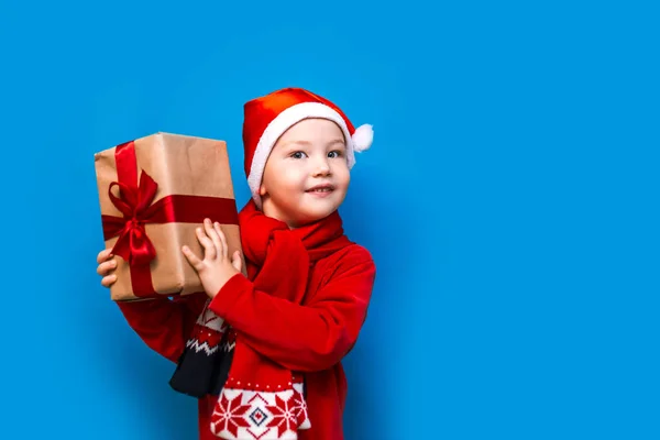 Niño en el sombrero de Santa con acogedora caja de regalo y cinta roja quieren saber lo que dentro de la caja, poner a su oído —  Fotos de Stock