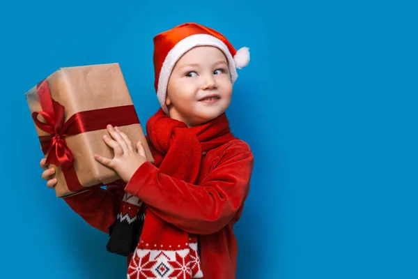 Niño en el sombrero de Santa con acogedora caja de regalo y cinta roja quieren saber lo que dentro de la caja, poner a su oído —  Fotos de Stock