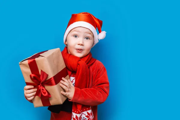 En Navidad. niño con Santa Claus Sombrero celebración de regalo de Navidad con cinta roja y wery cara sorprendida con la boca abierta —  Fotos de Stock