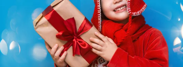 Niño en el sombrero de Santa con acogedora caja de regalo y cinta roja quieren saber lo que dentro de la caja, poner a su oído —  Fotos de Stock