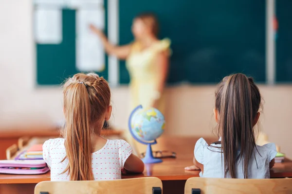 Två små skolflickor sitter vid ett skrivbord i en skolklass och lyssnar noga på läraren — Stockfoto