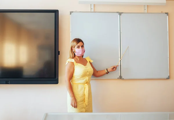 Young teacher in a medical mask holding a pointer in her hand and points to a white board, back to school, modern education, training during the epidemic.