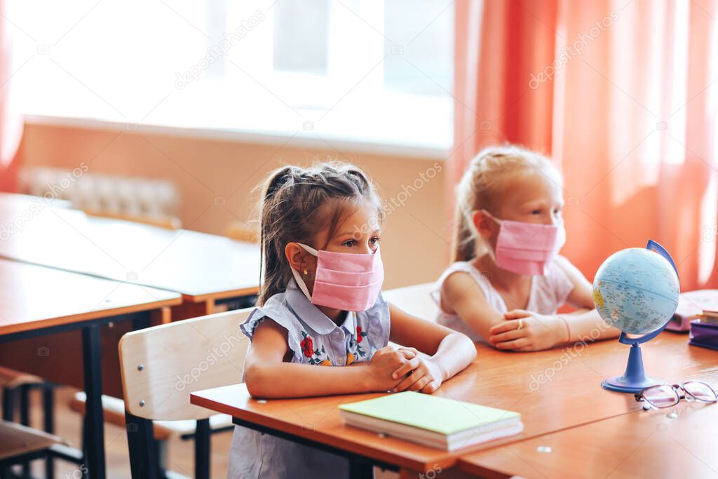 Two little schoolgirls sit at a desk in a school class in medical masks and carefully listen to the teacher