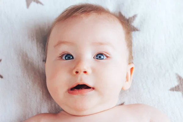 Фото ребенка с эмоцией удивления, новорожденного ребенка с голубыми глазами и светлыми волосами, лежащего на светлом одеяле с коричневыми звездами — стоковое фото