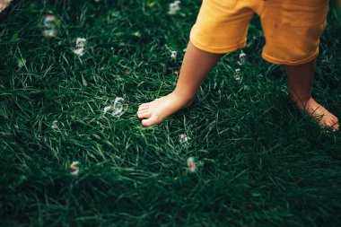 Yeşil bir çimenlikte çıplak ayakla duran küçük bir çocuk, bir çocuk ayaklarıyla sabun köpüklerini eziyor.