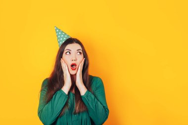 Şaşırmış yüzlü bir kadın, sarı arka planda elleri yeşil bir elbiseyle yüzünü tutuyor.