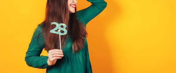 Красива жінка святкує свій день народження на жовтому тлі в зеленому платті, а на її голові святкову шапку тримає номер 28 в руці — стокове фото