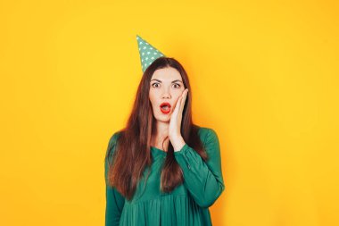 Şaşırmış yüzlü bir kadın, sarı arka planda elleri yeşil bir elbiseyle yüzünü tutuyor.