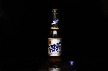 Front view of San Miguel Light beer with silver and blue label with the lid next to it in a black marble background. clipart