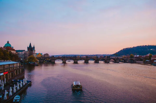 Gün batımında Vltava Nehri 'ni geçen bir tekne ve gökyüzünden yansıyan mor ile pembe renklerin gradyanı..
