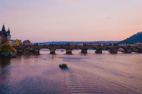 Gün batımında Vltava Nehri 'ni geçen bir tekne ve gökyüzünden yansıyan mor ile pembe renklerin gradyanı..