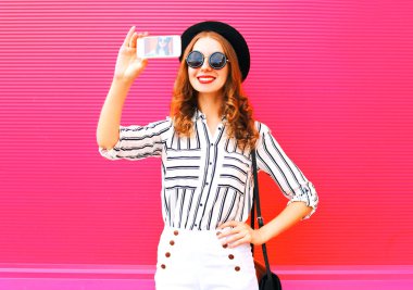 Smartphone giyme kendi kendine portre siyah şapka beyaz pantolon pembe renkli arka plan üzerinde çekim güzel kadın modeli