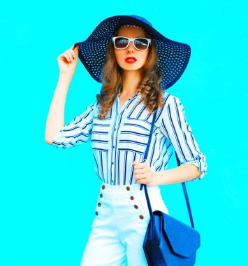 Şehirde poz renkli mavi arka plan üzerinde bir hasır şapka, beyaz pantolon ve el çantası debriyaj giyen zarif genç ve güzel kadın