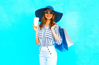 Şehirde poz renkli mavi arka plan üzerinde beyaz bir alışveriş torbaları, hasır şapka giyen kahve fincanı ile zarif kadın pantolon