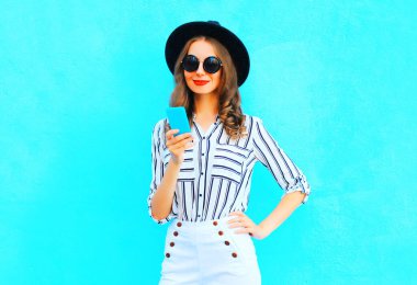 Zarif kız şehirde mavi renkli arka plan üzerinde siyah bir şapka ve çanta debriyaj giyen bir akıllı telefon kullanıyor