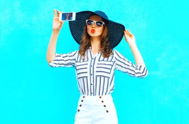 Güzel kız bir saman yaz şapka Smartphone'da mavi renkli arka plan üzerinde beyaz pantolon bir resim alıyor