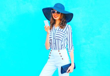 Mutlu kız bir hasır şapka giymiş bir akıllı telefon kullanıyor, beyaz bir el çantası debriyaj ile mavi renkli arka plan üzerinde pantolon 