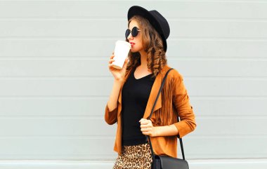 Genç kadın bir retro şık şapka, güneş gözlüğü, kahverengi ceket ve siyah çanta gri arka plan üzerinde giyen Kupası Kahve içer