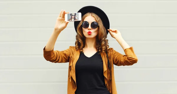 クールな女の子モデル都市の灰色の背景の上身に着けているレトロなエレガントな帽子 サングラス 茶色ジャケット 巻き毛を持つハンドバッグのスマート フォンで写真画像自画像を撮影 — ストック写真