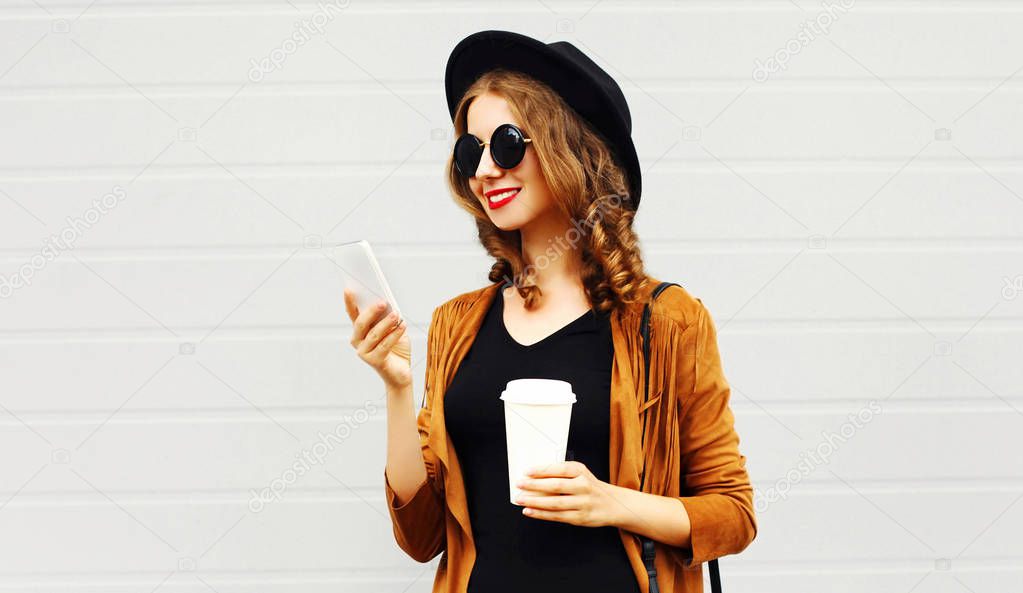 Beautiful cool smiling young woman with coffee cup using smartphone walking in city, happy female model wearing elegant hat, brown jacket outdoors over grey background
