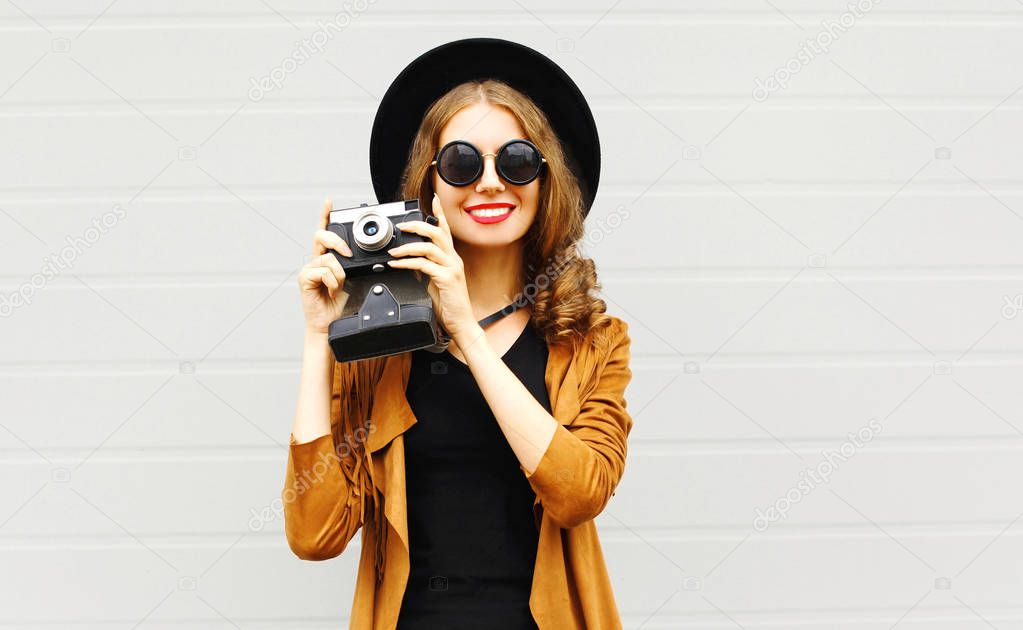 Happy cool young woman model with retro film camera wearing a elegant hat, brown jacket outdoors over city grey background