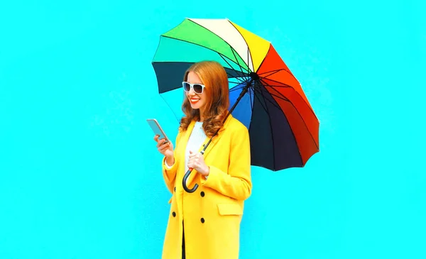 スマート フォンで幸せな笑みを浮かべて女をファッションが青の背景に黄色のコート ジャケットのカラフルな傘を保持します — ストック写真