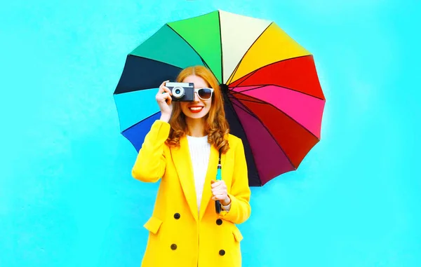 女性は笑みを浮かべて幸せなレトロなフィルム カメラで画像青の背景に黄色のコート ジャケットのカラフルな傘を保持します — ストック写真