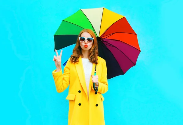 肖像画のクールな女の子は 青の背景に黄色のコート ジャケットのカラフルな傘と空気キスを送信します — ストック写真