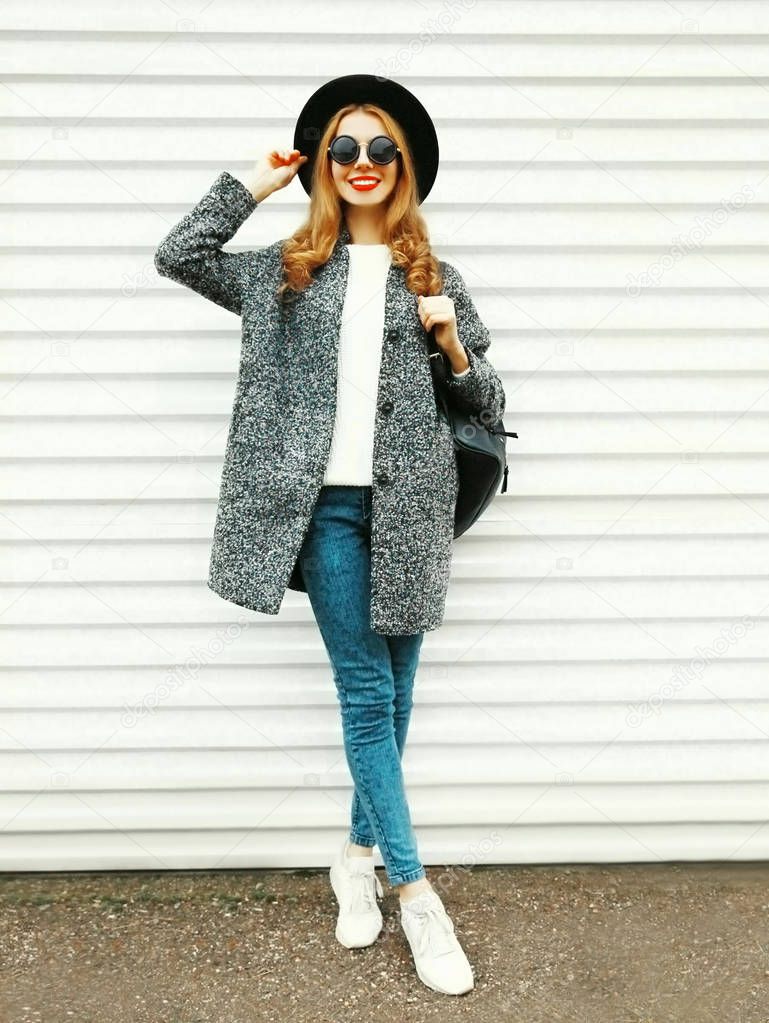 Fashion full-length woman in gray coat, black round hat posing on white wall background on street city