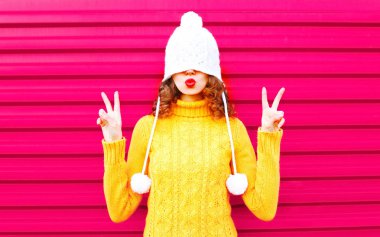 Pembe arka plan üzerinde giyen renkli örgü şapka, sarı kazak öp hava serin kız kırmızı dudaklar üfleme yapar