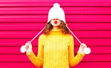 Pembe arka plan üzerinde giyen renkli örgü şapka, sarı kazak öp hava serin kız kırmızı dudaklar üfleme yapar