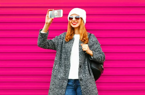 コート ジャケット ニット帽子 バックパック クールなヒップのカラフルなピンクの壁の背景に楽しんで身に着けているスマート フォンで Selfie を取って陽気な若い笑顔の女性 — ストック写真