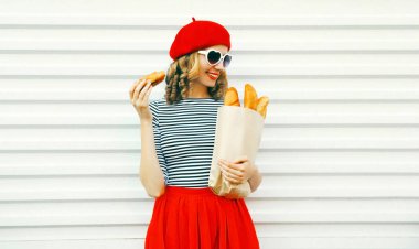 Kırmızı bere kruvasan, beyaz duvar arka plan üzerinde uzun beyaz ekmek baget ile kağıt torba tutarak giyen çok gülümseyen genç kadın