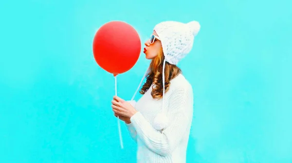 Happy woman kisses red air balloon in white knitted hat on blue