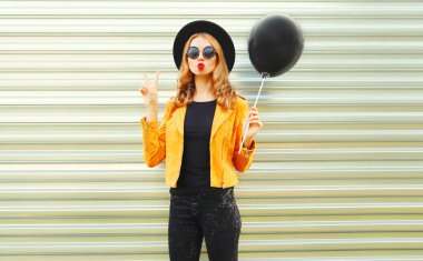 Kırmızı dudaklar üfleme poz mutlu serin kız yuvarlak şapka, sarı ceket metal duvar arka plan üzerinde siyah hava balonu holding hava öpücük gönderir