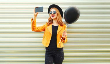 Selfie fotoğrafını smartphone holding siyah helyum hava balonu yuvarlak şapkalı, sarı ceket tarafından metal duvar arka plan üzerinde mutlu gülümseyen kadın