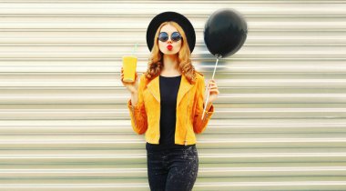 Sahip şık genç kadın kırmızı dudaklar gönderir hava üfleme eğlenceli öpücük siyah hava balonu, meyve suyu yuvarlak şapkalı, metal duvar zemin üzerine sarı ceket bir holding