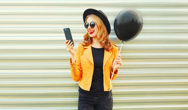 Portrait happy pretty smiling woman with phone holding black helium air balloon in round hat, yellow jacket on metal wall background