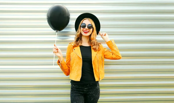 丸い帽子 金属製の壁の背景に黄色のジャケットに黒のヘリウム気球を保持している肖像画幸せ笑顔の女性 — ストック写真
