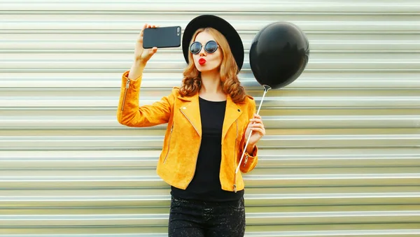 かなりクールな丸い帽子 金属製の壁の背景に黄色のジャケットに黒のヘリウム気球を保持している赤い唇を吹いてのスマート フォンで Selfie 写真を撮る女の子 — ストック写真