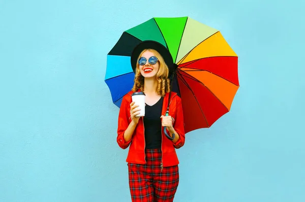 Портрет улыбающейся женщины с красочным зонтиком, чашкой горячего кофе, в красной куртке, черной шляпе на фоне синей стены — стоковое фото