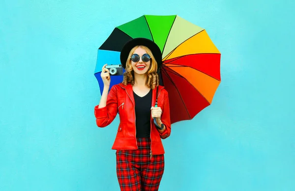 Szczęśliwy uśmiechnięta kobieta trzyma kolorowy parasol, retro aparat fotograficzny biorąc obraz w czerwonym płaszczu, czarny kapelusz na niebieskim tle ściany — Zdjęcie stockowe