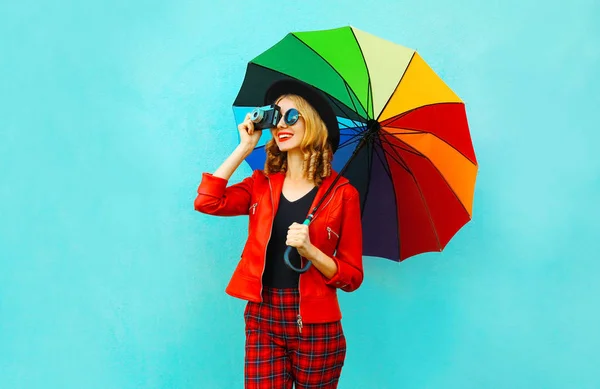 Happy smiling woman holding colorful umbrella, retro camera taking picture in red jacket, black hat on blue wall background — Stock Photo, Image