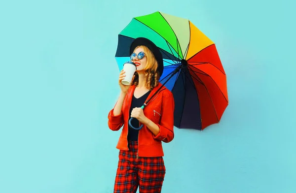 Молода жінка п'є каву і тримає барвисту парасольку, що йде в червоній куртці, чорний капелюх на фоні синьої стіни — стокове фото