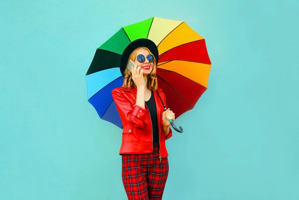 Портрет улыбается молодая женщина звонит по телефону с красочным зонтиком в красной куртке, черная шляпа на синем фоне стены — стоковое фото