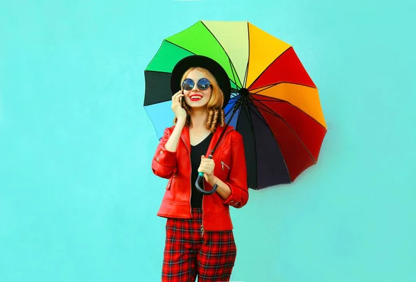 Retrato sorrindo jovem chamando no telefone com guarda-chuva colorido em casaco vermelho, chapéu preto no fundo da parede azul — Fotografia de Stock